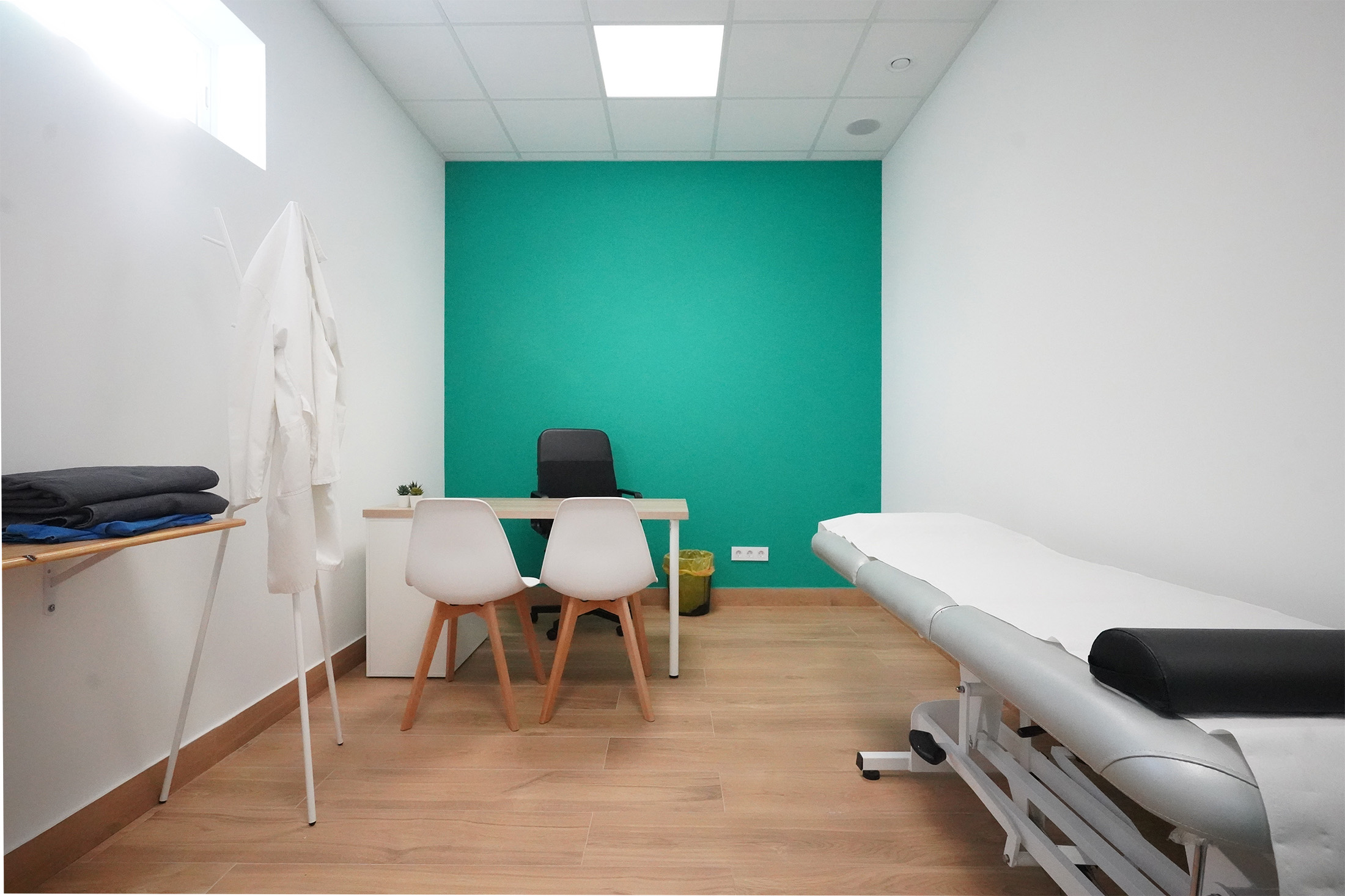 Consulta médica con escritorio, dos sillas de atención al paciente, camilla de exploración y pared de acento turquesa al fondo